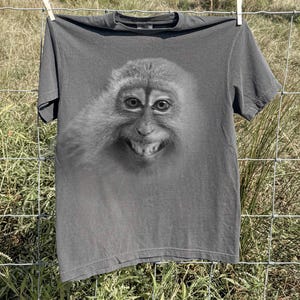 May include: Dark gray t-shirt featuring a close-up of a monkey's face with a wide grin. The monkey's fur is detailed in shades of gray, and the shirt is hanging on a clothesline. The shirt is made of a soft material.