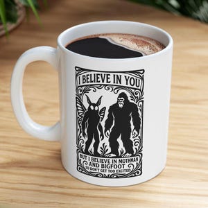 May include: White ceramic mug featuring a black graphic of Mothman and Bigfoot, with the text "I BELIEVE IN YOU" and "BUT I BELIEVE IN MOTHMAN AND BIGFOOT SO DON'T GET TOO EXCITED". The mug contains a dark liquid.