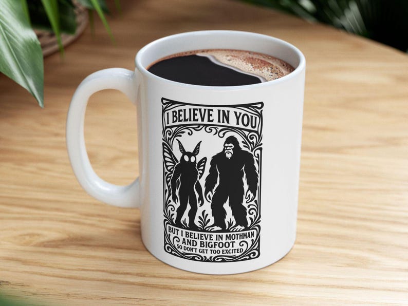May include: White ceramic mug featuring a black graphic of Mothman and Bigfoot, with the text "I BELIEVE IN YOU" and "BUT I BELIEVE IN MOTHMAN AND BIGFOOT SO DON'T GET TOO EXCITED". The mug contains a dark liquid.