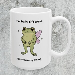 May include: White ceramic mug featuring a cartoon frog with fairy wings and the text "I'm built different (Like incorrectly I think)". The frog is green with a light green belly and pink wings. The mug has a large handle.