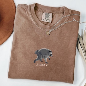 Pode incluir: Uma t-shirt castanha de manga curta com um gráfico de um guaxinim cinzento e o texto "JIMOTHY" impresso por baixo. A t-shirt tem um decote redondo e uma etiqueta da marca Comfort Colors no interior da gola indicando o tamanho S.
