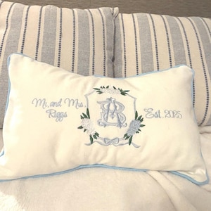 May include: Cream-colored decorative pillow with light blue embroidered details. Features a floral crest with the initials "RB", the text "Mr. and Mrs. Riggs", and "Est. 2025". The pillow has a light blue border.