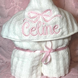 May include: A white hooded baby towel with the name "Celine" embroidered in pink script. The towel is tied with a pink ribbon and sits on a pink, bow-patterned surface.