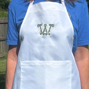 May include: A white apron featuring a large, embroidered green letter W in a decorative, swirling font centered on the chest. The apron includes a horizontal front pocket across the waist area and is secured with white neck and waist ties.