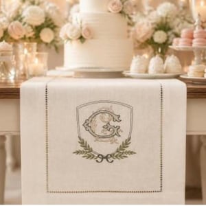 May include: A cream-colored table runner with a decorative embroidered crest featuring the letter "G" and laurel leaves. The runner is placed on a wooden table, with a tiered wedding cake, cupcakes, and macarons in the background.