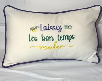 May include: A rectangular off-white throw pillow with dark purple piping along the edges. The front features embroidered text that reads Laissez les bon temps rouler in purple, green, and yellow, accompanied by a jester hat and a masquerade mask.