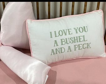 May include: A rectangular white pillow with light green embroidered text that reads I LOVE YOU A BUSHEL AND A PECK. The pillow features light pink piping along the edges and is positioned next to a soft, shiny light pink fabric bow accessory.