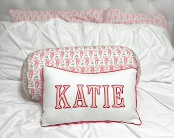 May include: A rectangular white throw pillow with a textured surface featuring the name KATIE embroidered in pink and orange thread, bordered by pink piping. Behind it sits a cylindrical bolster pillow with a pink and green monkey pattern.