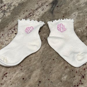 May include: A pair of white ankle socks featuring a ruffled top edge and a pink embroidered monogram on the side. The socks are made of a ribbed knit fabric and display the letters BGL in a decorative script font on each individual sock.