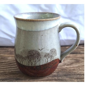 May include: A ceramic mug featuring a two-tone glaze with a cream-colored upper section and a textured, dark reddish-brown base. The cream portion displays a detailed brown etched illustration of a forest landscape with bare trees and birds.