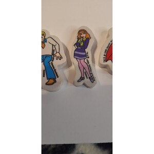 Puede incluir: Una colección de gomas de borrar vintage con personajes de dibujos animados. Una goma muestra a una mujer con un vestido morado y medias rosas. Las gomas son blancas con ilustraciones coloridas y texto.