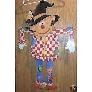 May include: A colorful, cartoonish scarecrow decoration with a black hat, a checkered red and white shirt, and blue pants. The scarecrow has a friendly face and is adorned with patches. A black crow sits on the hat. Perfect for fall or Halloween.