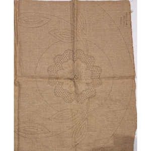 May include: A tan fabric with a floral embroidery pattern. The design features a large flower in the center, surrounded by a circular outline and several leaf shapes. The embroidery is done in a dark brown thread.