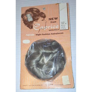 May include: A vintage hairpiece, encased in clear plastic, is displayed on a retro-style cardboard backing. The hairpiece is a blend of gray and light brown, with a curly texture. The packaging features the text "NEW from Corie Originals" and "Tainelle high-fashion hairpieces."