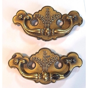 May include: Two matching brass drawer pulls featuring a decorative backplate with a central embossed wheat sheaf motif. Each pull has a curved, swinging handle attached to the scalloped backplate, which includes two circular mounting holes.