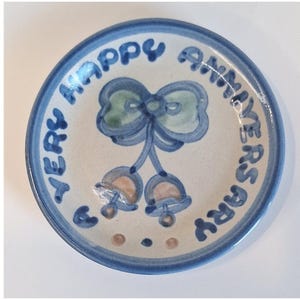 Puede incluir: Un plato de cerámica pequeño y redondo con un borde azul y las palabras "VERY HAPPY ANNIVERSARY" en letras azules. El centro presenta un lazo azul y dos diseños en forma de campana con acentos melocotón y verdes.