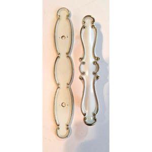 Peut inclure: Deux poignées de placard allongées avec une finition blanche lisse et des bords dorés métalliques. L'une est une plaque arrière plate avec deux trous de vis circulaires, l'autre est une poignée surélevée au design incurvé et orné.