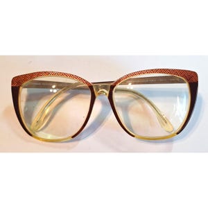 May include: Anne Klein eyeglasses featuring a cat-eye frame with a dark brown and amber gradient finish. The upper rim displays a distinct chevron pattern in a reddish-brown tone. The interior temple arm is marked with the text HANDMADE FRAME ITALY.