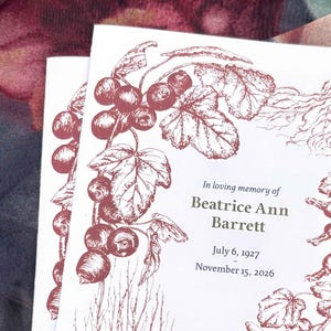 May include: A white memorial card featuring a dark red botanical illustration of currant berries and leaves framing the text: In loving memory of Beatrice Ann Barrett, July 6, 1927 - November 15, 2026. The design includes detailed line art.
