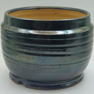 Puede incluir: Jardinera de cerámica hecha a mano con un esmalte verde azulado oscuro y textura acanalada horizontal. El interior es de color marrón claro. La jardinera tiene forma redondeada con un borde ligeramente ensanchado y una base pequeña.