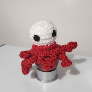 Peut inclure: Poupée pieuvre au crochet faite à la main, avec une tête blanche et un corps rouge. La poupée a de grands yeux sombres et est posée sur un petit récipient argenté. La pieuvre a huit tentacules rouges.