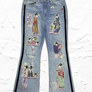 Puede incluir: Vaqueros de mezclilla de lavado claro con figuras de geishas bordadas y parches florales. Los vaqueros tienen rayas laterales negras y un pequeño dije dorado. Los detalles desgastados añaden un aspecto vintage a estos vaqueros únicos y artísticos.