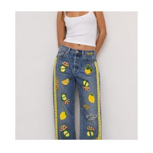 Puede incluir: Vaqueros azules con rayas laterales amarillas y parches con temática de limón. Los vaqueros tienen bragueta con botones, detalles desgastados y varios diseños de limones bordados. También se ve una camiseta sin mangas blanca.