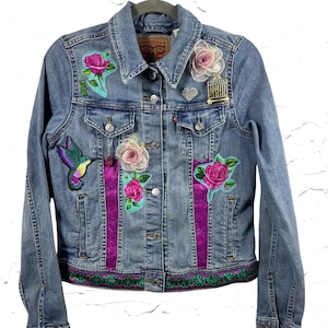 Könnte beinhalten: Hellblaue Jeansjacke mit aufgestickten Blumen- und Kolibri-Patches. Mit dekorativem Besatz am Saum und lila Bändern. Inklusive einer kleinen Vogelkäfig-Anstecknadel und einer silbernen Herz-Anstecknadel.
