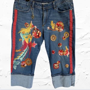 Könnte beinhalten: Blaue Denim-Capri-Jeans mit gestickten Blumen- und Vogelmotiven. Rote Tupfenstreifen verlaufen an den Seiten. Die Jeans hat einen umgeschlagenen Saum und einen dekorativen Anhänger am Gürtelschlaufen.
