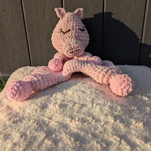 May include: A handmade, pink crocheted pig-shaped plush toy with closed eyes and a smiling expression. The pig has long, curved arms and is resting on a white, fluffy surface. The background is a dark gray wooden wall.