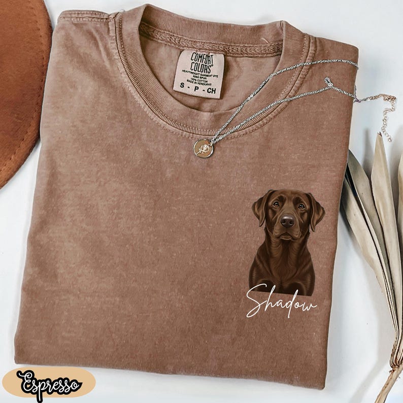 May include: Espresso-colored Comfort Colors t-shirt featuring a brown Labrador Retriever graphic with the name "Shadow" in white script. A silver necklace with a pendant rests on the shirt. The shirt has a crew neck.