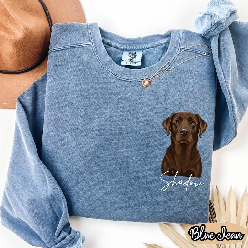 May include: A blue jean-colored sweatshirt featuring a brown Labrador Retriever graphic with the name "Shadow" in white script. The sweatshirt has a crew neck and long sleeves. The words "Blue Jean" are printed in a script font at the bottom.