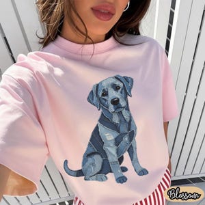 May include: A light pink t-shirt featuring a blue dog illustration made of denim patches. The dog is sitting with its tail curled. The word "Blossom" is printed in a cursive font on the bottom right corner of the shirt.