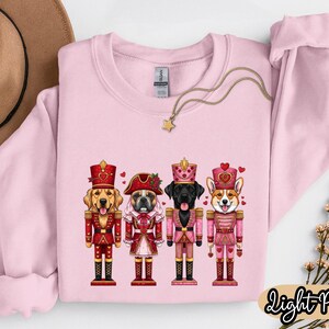 May include: Light pink sweatshirt featuring a graphic of four dogs dressed as nutcrackers. The dogs are a golden retriever, a French bulldog, a black Labrador, and a Corgi. Each dog is wearing a different hat and uniform. A gold necklace with a star pendant is draped across the sweatshirt.