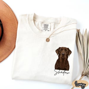 May include: Ivory-colored t-shirt featuring a brown Labrador Retriever illustration with the name "Shadow" in a script font. The shirt has a Comfort Colors tag and a gold necklace. The word "Ivory" is written in a script font on the bottom right.
