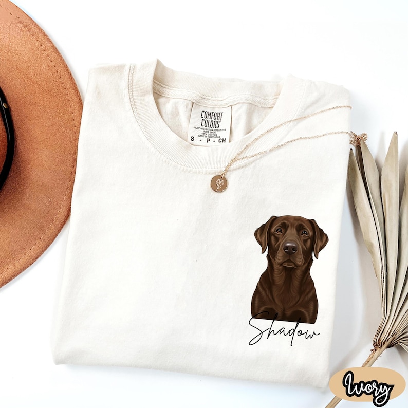 May include: Ivory-colored t-shirt featuring a brown Labrador Retriever illustration with the name "Shadow" in a script font. The shirt has a Comfort Colors tag and a gold necklace. The word "Ivory" is written in a script font on the bottom right.