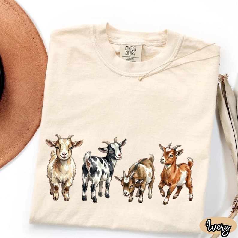 May include: Ivory-colored t-shirt featuring a graphic of four goats in various colors. The goats are depicted in a cartoon style, with detailed fur and horns. The shirt has a crew neck and short sleeves. The brand name "Comfort Colors" is visible on the tag.