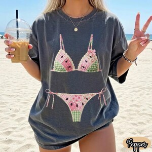 May include: A charcoal gray short-sleeve t-shirt featuring a graphic print of a two-piece bikini with a patchwork pattern of pink, green, and white colors, including watermelon seed motifs and a small pink bow on the top piece.