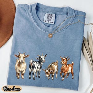 May include: A blue jean colored Comfort Colors t-shirt featuring a graphic of four goats in various colors. The shirt has a crew neck and short sleeves. A gold necklace with a pendant is draped on the shirt. The words "Blue Jean" are printed on the bottom.