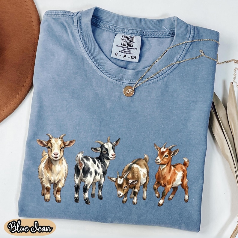 May include: A blue jean colored Comfort Colors t-shirt featuring a graphic of four goats in various colors. The shirt has a crew neck and short sleeves. A gold necklace with a pendant is draped on the shirt. The words "Blue Jean" are printed on the bottom.