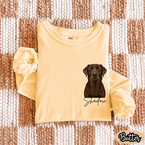 May include: A long-sleeved, butter-yellow t-shirt featuring a detailed illustration of a chocolate Labrador Retriever. The dog's name, "Shadow," is written in cursive below the image. The word "Butter" is printed in a rounded font.