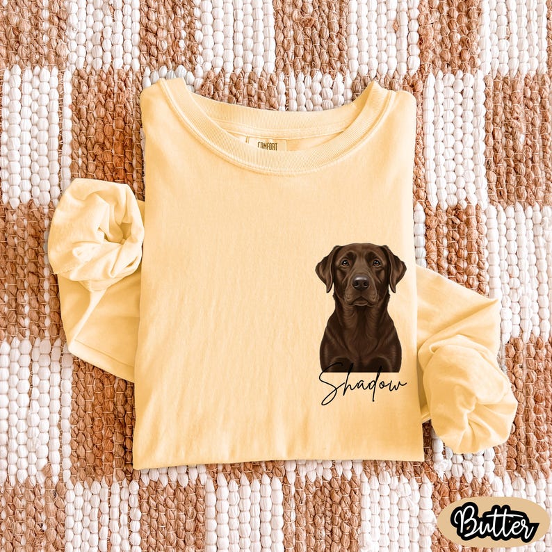 May include: A long-sleeved, butter-yellow t-shirt featuring a detailed illustration of a chocolate Labrador Retriever. The dog's name, "Shadow," is written in cursive below the image. The word "Butter" is printed in a rounded font.