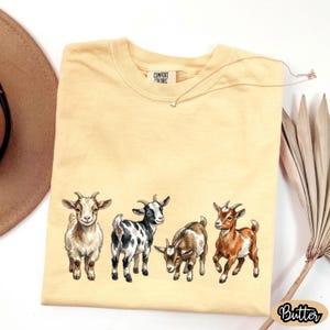 May include: A light yellow t-shirt featuring a row of five illustrated goats in various colors and poses. The shirt has the text "COMFORT COLORS" on the tag and the word "Butter" in a cursive font on the bottom right.