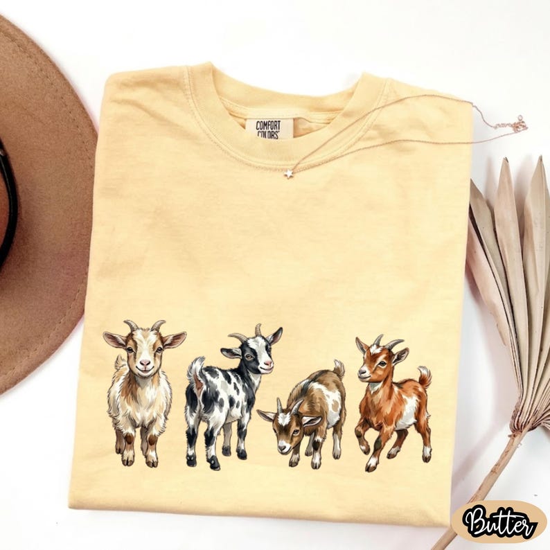 May include: A light yellow t-shirt featuring a row of five illustrated goats in various colors and poses. The shirt has the text "COMFORT COLORS" on the tag and the word "Butter" in a cursive font on the bottom right.