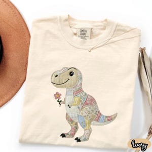 May include: Ivory-colored t-shirt featuring a smiling dinosaur holding a pink flower. The dinosaur is made of patchwork fabric in various floral and patterned designs. The shirt has a Comfort Colors tag.