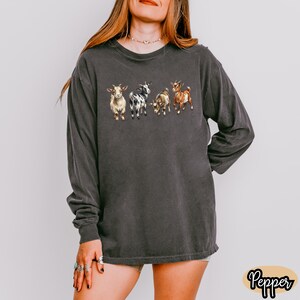 May include: Dark gray long-sleeve shirt featuring a watercolor illustration of four goats. The goats are in various colors, including tan, black and white, and brown. The shirt has a crew neck and long sleeves.
