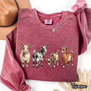 May include: Crimson-colored sweatshirt featuring a watercolor illustration of four goats. The goats are in various colors, including white, brown, and black. The sweatshirt has a crew neck and long sleeves. The word "Crimson" is written in a cursive font on the bottom right.
