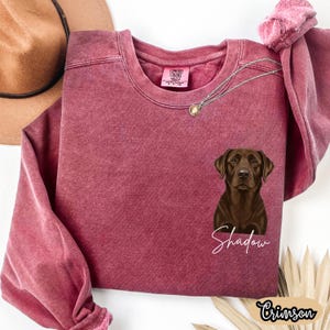 May include: Crimson-colored sweatshirt with a brown Labrador Retriever graphic and the name "Shadow" in white script. The sweatshirt has a crew neck and long sleeves. A gold necklace is draped across the front.