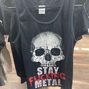 May include: Black tank top featuring a detailed skull graphic. The text "STAY FUCKING METAL" is printed in red and white below the skull. The tank top is hanging on a black hanger.