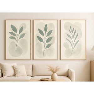 Puede incluir: Tres impresiones artísticas botánicas enmarcadas. Cada impresión presenta un diseño de hojas verdes sobre un fondo crema con un lavado de acuarela verde claro. Los marcos son de color madera clara. Las impresiones se exhiben sobre un sofá color crema.
